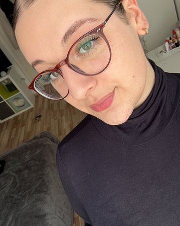 Eine Person mit kurzen, glatten Haaren und Brille trägt ein schwarzes Oberteil. Sie steht in einem hellen Raum mit Möbeln im Hintergrund.