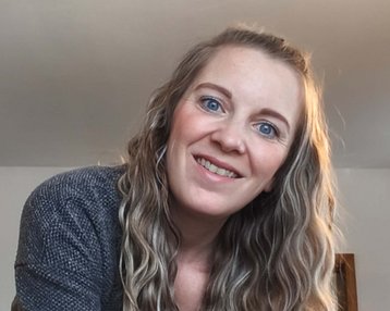 Eine Frau mit langen, lockigen Haaren lächelt in die Kamera, während sie sich leicht nach vorne beugt. Sie trägt einen grauen Pullover.