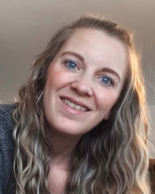 Eine Frau mit langen, lockigen Haaren lächelt in die Kamera, während sie sich leicht nach vorne beugt. Sie trägt einen grauen Pullover.