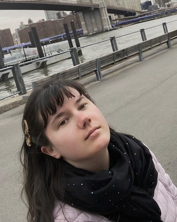 Eine Person mit langen, dunklen Haaren und einem schwarzen Schal steht am Wasser, mit der Brooklyn Bridge und der Skyline im Hintergrund.