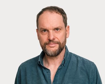 Ein Mann mit einem Bart und kurzen Haaren trägt ein blaues Hemd. Er blickt direkt in die Kamera vor einem neutralen Hintergrund.