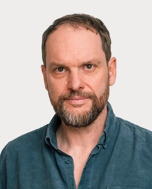 Ein Mann mit einem Bart und kurzen Haaren trägt ein blaues Hemd. Er blickt direkt in die Kamera vor einem neutralen Hintergrund.