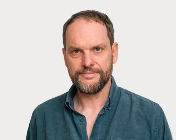 Ein Mann mit einem Bart und kurzen Haaren trägt ein blaues Hemd. Er blickt direkt in die Kamera vor einem neutralen Hintergrund.