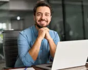 Ein lächelnder Mann sitzt an einem Schreibtisch vor einem Laptop in einem modernen Büro.