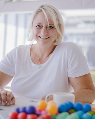 Eine lächelnde Frau mit blonden Haaren sitzt an einem Tisch mit einem Laptop und einer Tasse. Vor ihr liegt eine bunte Ansammlung von kleinen Kugeln.