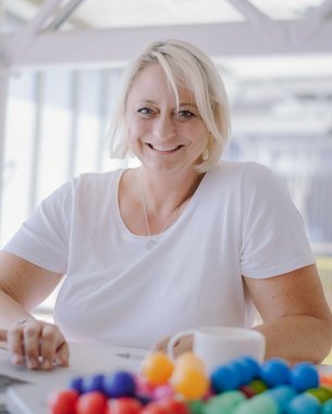 Eine lächelnde Frau mit blonden Haaren sitzt an einem Tisch mit einem Laptop und einer Tasse. Vor ihr liegt eine bunte Ansammlung von kleinen Kugeln.