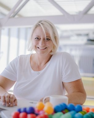 Marion Krüger-Altena, Studentin Psychologie im Online-Abendstudium (B.Sc.) | SRH Fernhochschule Eine lächelnde Frau mit blonden Haaren sitzt an einem Tisch mit einem Laptop und einer Tasse. Vor ihr liegt eine bunte Ansammlung von kleinen Kugeln.