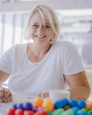 Eine lächelnde Frau mit blonden Haaren sitzt an einem Tisch mit einem Laptop und einer Tasse. Vor ihr liegt eine bunte Ansammlung von kleinen Kugeln.