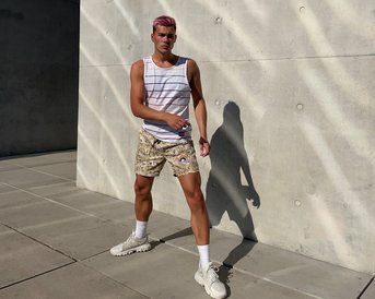 Ein junger Mann mit pinken Haaren trägt ein gestreiftes Tanktop und gemusterte Shorts. Er steht auf einem gepflasterten Boden vor einer grauen Wand und hat eine entspannte Pose.