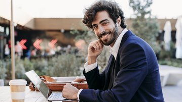 Ein Mann mit lockigem Haar sitzt an einem Tisch im Freien, lächelt und schaut zur Kamera. Vor ihm stehen ein Laptop, eine Kaffeetasse und einige Unterlagen.