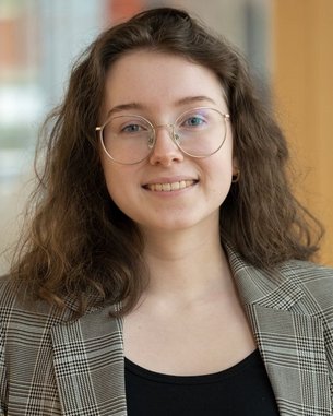 Junge Frau mit lockigem Haar und Brille, die ein kariertes Blazer-Jackett trägt. Sie steht in einem modernen Innenraum mit Holz- und Glaselementen.