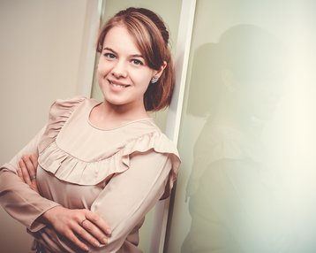 Eine junge Frau mit einem lächelnden Gesicht steht mit verschränkten Armen vor einer Wand. Sie trägt eine blusige, rüschige Bluse und hat eine elegante Frisur. Ihr Schatten ist leicht im Hintergrund sichtbar.