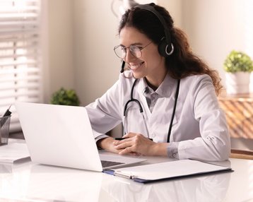 Eine Ärztin mit Headset sitzt an einem Tisch und lächelt, während sie auf einen Laptop schaut. Auf dem Tisch liegt ein Notizblock. Pflanzen sind im Hintergrund sichtbar.