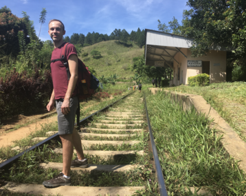 Ein Mann steht auf einem Bahngleis, umgeben von üppigem Grün und einer kleinen Station im Hintergrund. Er trägt einen Rucksack und blickt zur Kamera.