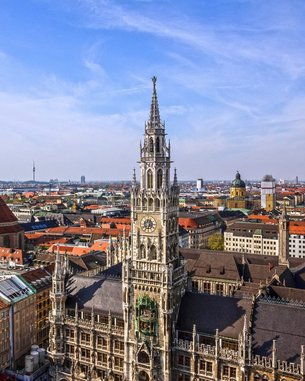 Moderne Gebäude mit großen Fenstern und einem überdachten Außenbereich. Tische und Stühle unter grünen Sonnenschirmen sind im Vordergrund sichtbar.