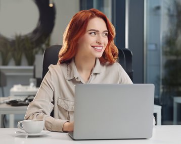 Eine lächelnde Frau mit roten Haaren sitzt an einem Schreibtisch vor einem Laptop. Auf dem Tisch steht eine Tasse Kaffee. Der Hintergrund zeigt eine moderne Büroumgebung.