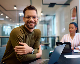 Ein lächelnder Mann mit Brille sitzt an einem Tisch mit einem Laptop, während eine Frau im Hintergrund an einem Tablet arbeitet.