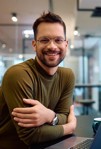 Ein lächelnder Mann mit Brille sitzt an einem Tisch mit einem Laptop, während eine Frau im Hintergrund an einem Tablet arbeitet.