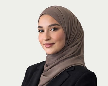 Eine junge Frau mit einem braunen Hijab und einem schwarzen Blazer, die in die Kamera lächelt. Der Hintergrund ist neutral gehalten.