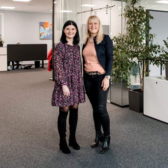 Zwei Frauen stehen in einem modernen Büro mit Pflanzen und Schreibtischen. Die Frau links trägt ein blumiges Kleid, die Frau rechts eine Bluse und eine Jacke. Im Hintergrund sind weitere Büromöbel sichtbar.