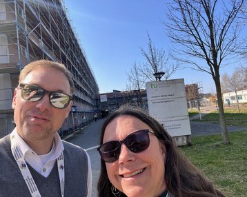 Zwei Personen posieren vor einem Gebäude mit Gerüst, das Schild der Technischen Universität ist im Hintergrund sichtbar.