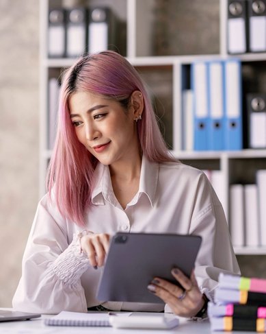Eine Frau mit pinken Haaren sitzt an einem Schreibtisch, nutzt ein Tablet und schaut auf einen Laptop. Im Hintergrund sind Aktenordner und eine kleine Pflanze sichtbar.