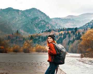 Eine Frau mit Rucksack steht am Ufer eines Flusses, umgeben von herbstlichen Bergen und Bäumen. Sie trägt einen roten Pullover und eine Mütze, während sie in die Kamera lächelt.