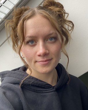 Junge Frau mit blonden, lockigen Haaren, die zu zwei Duttfrisuren gestylt sind. Sie trägt einen schwarzen Hoodie und schaut direkt in die Kamera.