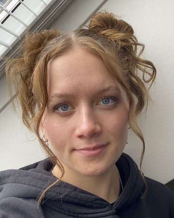 Junge Frau mit blonden, lockigen Haaren, die zu zwei Duttfrisuren gestylt sind. Sie trägt einen schwarzen Hoodie und schaut direkt in die Kamera.