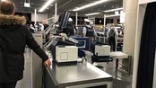 Sicherheitskontrolle am Flughafen mit mehreren Personen, darunter Polizisten. Ein Reisender steht an einem Kontrolltisch, während andere Passagiere im Hintergrund warten.