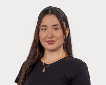Eine junge Frau mit langen, dunklen Haaren trägt ein schwarzes T-Shirt und eine goldene Halskette. Sie schaut direkt in die Kamera.