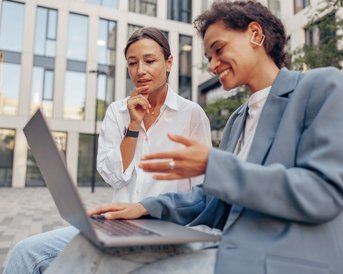 Zwei Frauen sitzen im Freien und schauen auf einen Laptop. Eine Frau zeigt auf den Bildschirm, während die andere nachdenklich wirkt. Beide tragen moderne Kleidung und sind in einem urbanen Umfeld.