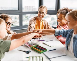 Eine Gruppe von fünf Kindern und einer Lehrerin sitzt an einem Tisch und legt ihre Hände übereinander. Auf dem Tisch liegen Schreibmaterialien und Notizblöcke. Die Atmosphäre wirkt kooperativ und freundlich.
