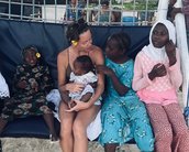 Eine Frau sitzt auf einer Bank am Strand und hält ein Baby in den Armen. Um sie herum sind mehrere Mädchen in bunten Kleidern, die miteinander sprechen und lächeln.