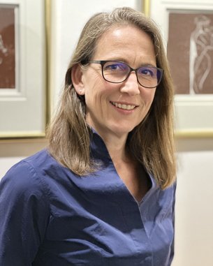 Eine Frau mit langen, glatten Haaren und einer blauen Bluse steht lächelnd vor gerahmten Kunstwerken. Sie trägt eine Brille und hat einen freundlichen Ausdruck.
