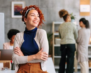 Eine lächelnde Frau mit lockigem, rotem Haar und einem Stirnband steht in einem modernen Büro. Im Hintergrund sind zwei weitere Frauen zu sehen, die miteinander sprechen.