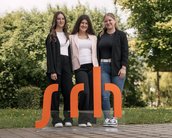Drei Frauen stehen vor einem orangefarbenen Schild mit der Aufschrift "sdh". Sie tragen formelle Kleidung und lächeln in die Kamera.
