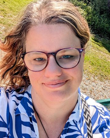 Frau mit Brille und blauer Bluse mit Blattmuster, lächelt in die Kamera, im Freien mit grünem Hintergrund.