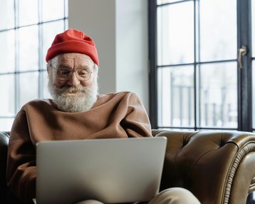 Ein älterer Mann mit Brille und rotem Hut sitzt auf einem Ledersofa und schaut auf einen Laptop. Er trägt einen braunen Pullover und hat einen grauen Bart.