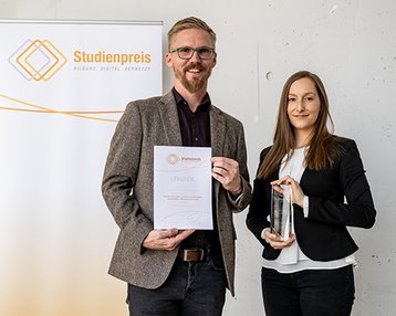 Zwei Personen halten eine Auszeichnung und ein Zertifikat. Im Hintergrund steht ein Banner mit dem Schriftzug "Studienpreis". Die Person links trägt einen grauen Blazer und die Person rechts ein schwarzes Jackett.