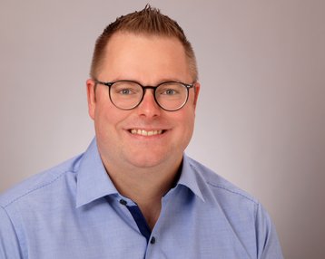 Ein lächelnder Mann mit Brille trägt ein hellblaues Hemd. Der Hintergrund ist neutral und hell.