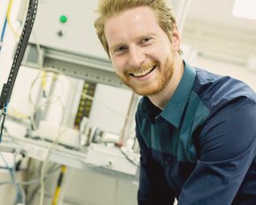 Ein lächelnder Mann mit blonden Haaren trägt ein blaues Hemd und arbeitet an einer technischen Apparatur in einem Labor. Kabel und Geräte sind im Hintergrund sichtbar.