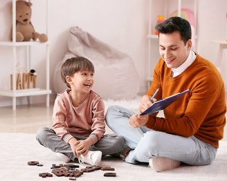 Ein Erwachsener und ein Kind sitzen auf dem Boden und spielen mit Buchstaben aus Holz. Der Erwachsene schreibt in ein Notizbuch, während das Kind lächelt und aufmerksam zuschaut.
