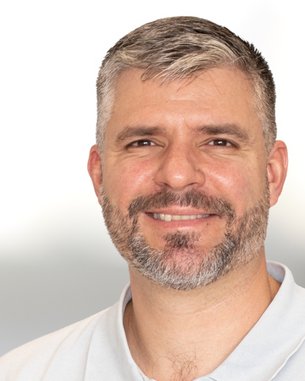Ein Mann mit grauen Haaren und Bart lächelt freundlich. Er trägt ein helles Polohemd und hat einen neutralen Hintergrund.