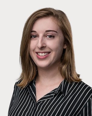 Eine junge Frau mit langen, lockigen Haaren trägt ein gestreiftes Hemd und lächelt in die Kamera. Der Hintergrund ist neutral.