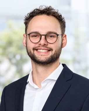 Ein junger Mann mit Brille und Bart lächelt, trägt einen Anzug und ein Hemd. Der Hintergrund zeigt unscharfe, helle Farben.