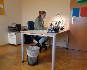 Eine Person sitzt an einem Schreibtisch in einem Büro, arbeitet am Computer und hat verschiedene Unterlagen sowie Büromaterialien auf dem Tisch. Ein Papierkorb steht neben dem Tisch.