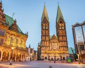 Der Bremer Dom mit seinen markanten Türmen und der großen Rosette steht im Vordergrund, umgeben von historischen Gebäuden und moderner Architektur. Die Szene ist bei Dämmerung beleuchtet.