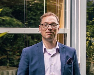 Ein Mann in einem Anzug steht entspannt vor einer Glaswand, umgeben von Pflanzen. Er trägt eine Brille und hat die Hände in den Taschen.