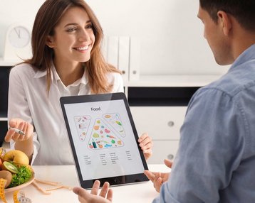 Eine lächelnde Frau mit lockigem Haar sitzt an einer Küchentheke, schreibt in ein Notizbuch und umgeben von frischem Gemüse und Obst.